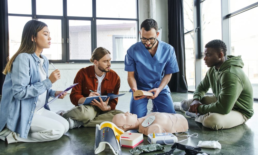 medical instructor showing wound care simulator to multiethnic team with clipboards near CPR manikin and medical equipment in training room, effective life-saving skills concept